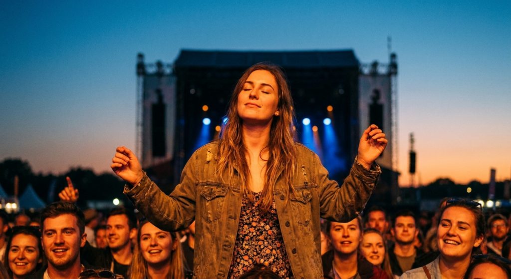 Young woman in a festival crowd at dusk, eyes closed and arms raised, absorbed in live music at a large outdoor UK music festival