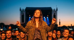 Young woman in a festival crowd at dusk, eyes closed and arms raised, absorbed in live music at a large outdoor UK music festival